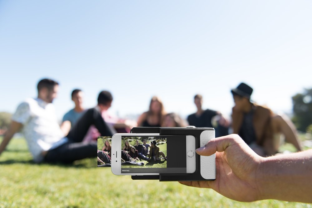 Une main tient une imprimante photo Prynt sur un smartphone, qui est en train d'imprimer la photo d'un groupe de jeunes gens assis dans l'herbe en extérieur
