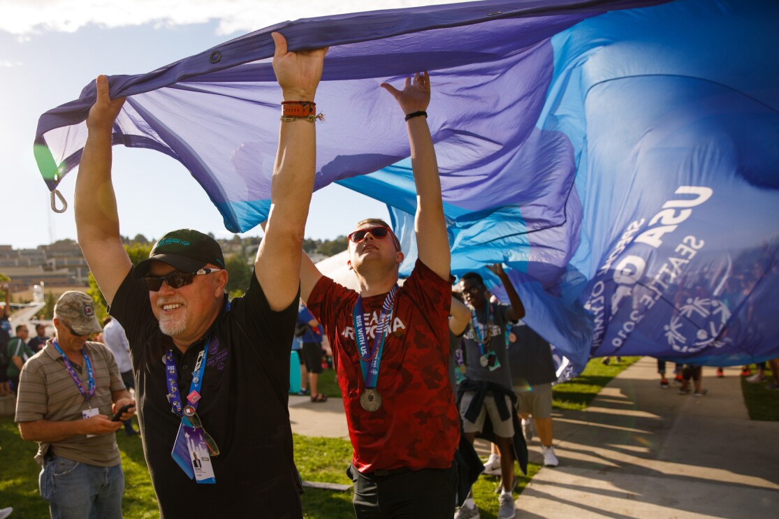Male athletes seen carrying the ceremonial Special Olympics flag. Athletes are seen holding the flag on the outer edges, while carrying it toward a destination. The athlete in the foreground is smiling while holding the flag with both hands. The second athlete is looking upward while holding onto the flag, while other athletes are seen behind him. The flag extends behind the first two athletes by several feet. Event attendees are watching, in the background.