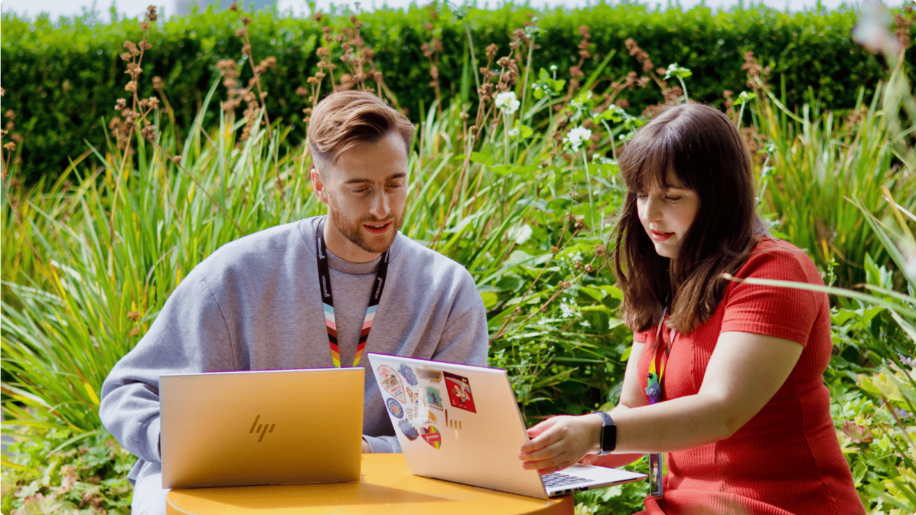 Two colleagues working on laptops in an outdoor settings, with greenery behind them