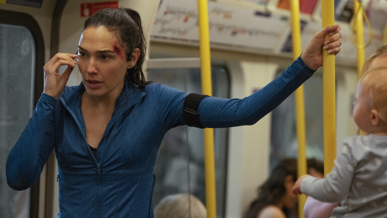 Woman in running gear stands on a train while on the phone, looking tense and with a cut at her brow
