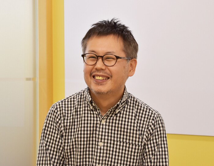 白い壁の前で笑顔を見せる男性。黒ぶち眼鏡で、黒と白のチェックのシャツを着用。