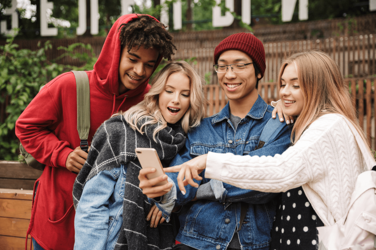 Four teenagers gather around and look in excitement at one of the teen's phone.