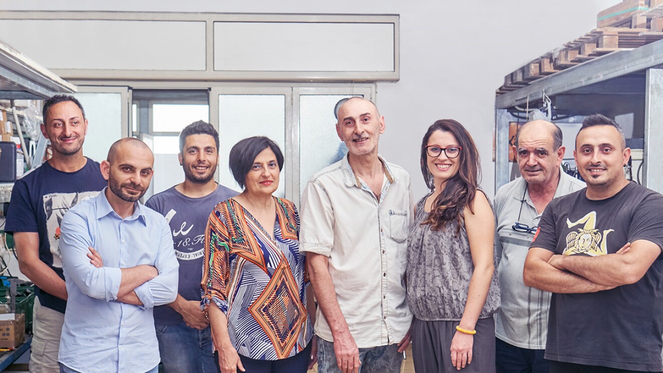 Foto del team di Kamira, formato da 8 persone in piedi, 6 uomini e 2 donne, tutti sorridenti, inclusi il fondatore Nino Santoro e la sua famiglia.