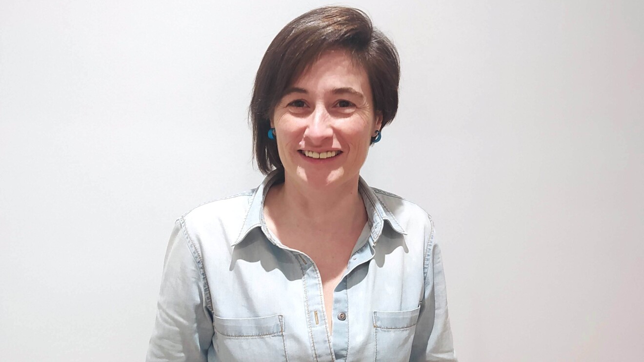 Marta Casasayas. Detrás un fondo blanco de pared. Marta aparece en un plano hasta los muslos. Lleva una camisa tejana azul clara de manga larga y unos jeans azul oscuro. Tiene el pelo corto y la parte izquierda más larga que la derecha. Está sonriendo y lleva unos pendientes que osn unos aros de color azul.