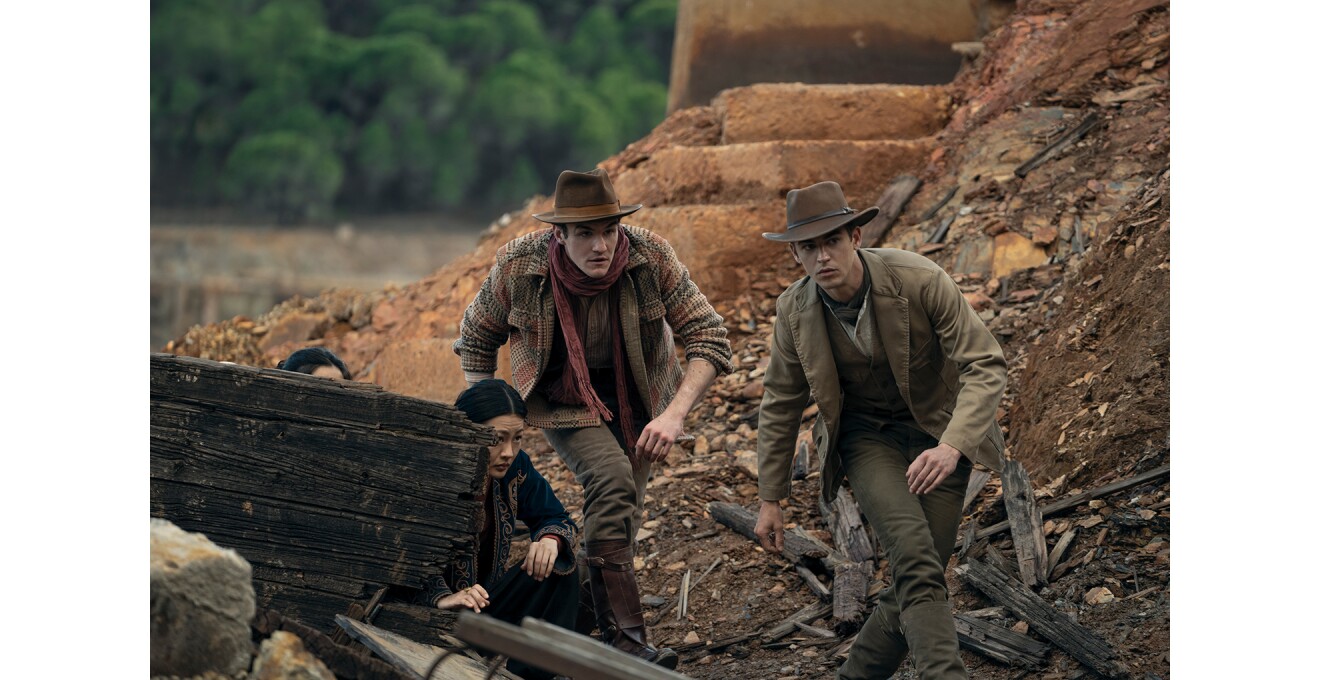 工事現場のような屋外で身をかがめて移動する2人の男性。ともに茶色い服装でカウボーイのような帽子をかぶっている。2人の左側にも人物が2人木の板の陰に隠れている。