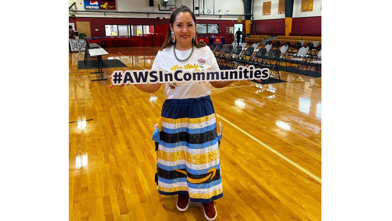 An image of a woman standing in a school gym holding a sign that says "#AWSincommunities"