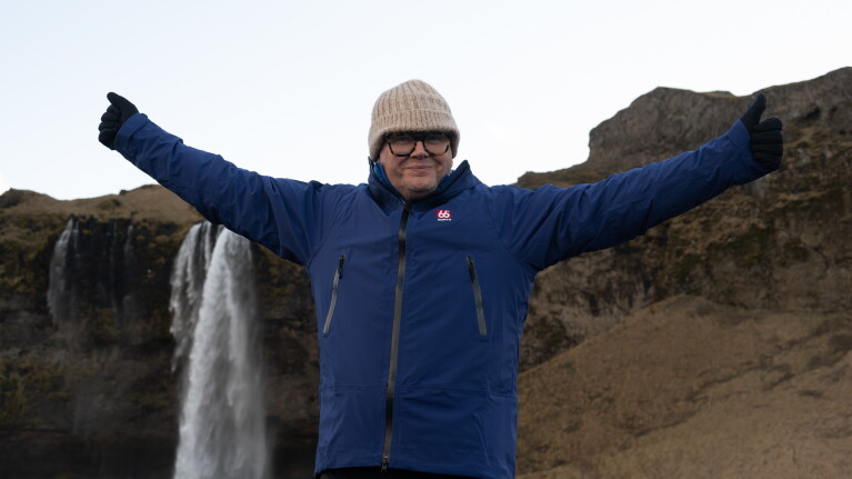 Alan Carr smiles with arms outstretched near rocky cliffs and waterfall