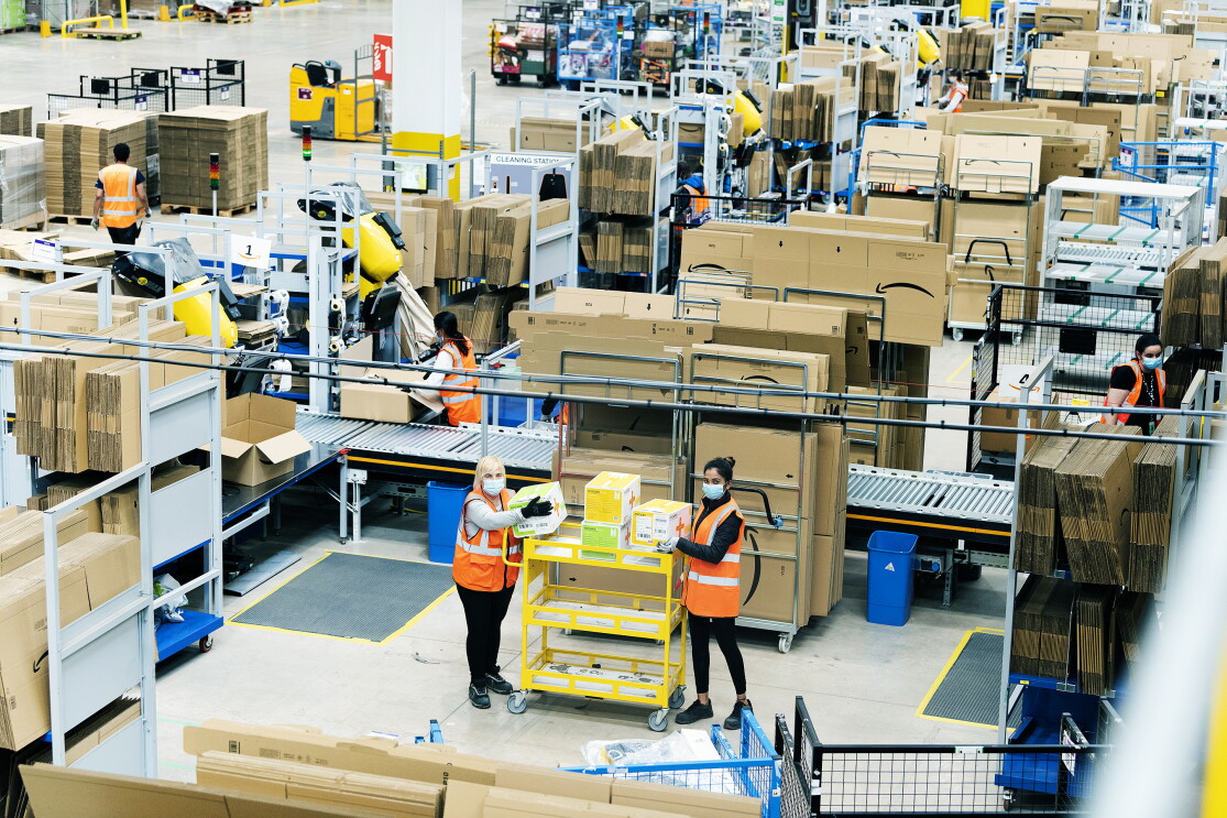 Wide shot of Amazon fulfilment centre staff stacking a yellow cart with medical supplies in the Coalville Fulfilment centre to provide supplies to the relevant government centres. They are wearing protective gloves and face masks while maintaining a social distance. 