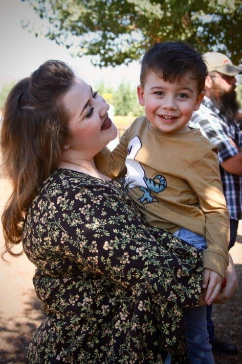 An image of a mother holding her son while looking up at him and smiling. Her son is also smiling and they are outside at a family gathering.