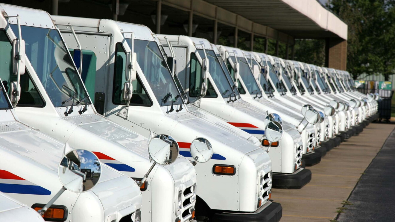 More than a dozen United States Postal Services delivery vehicles lined up under the canopy of a Postal Service building.