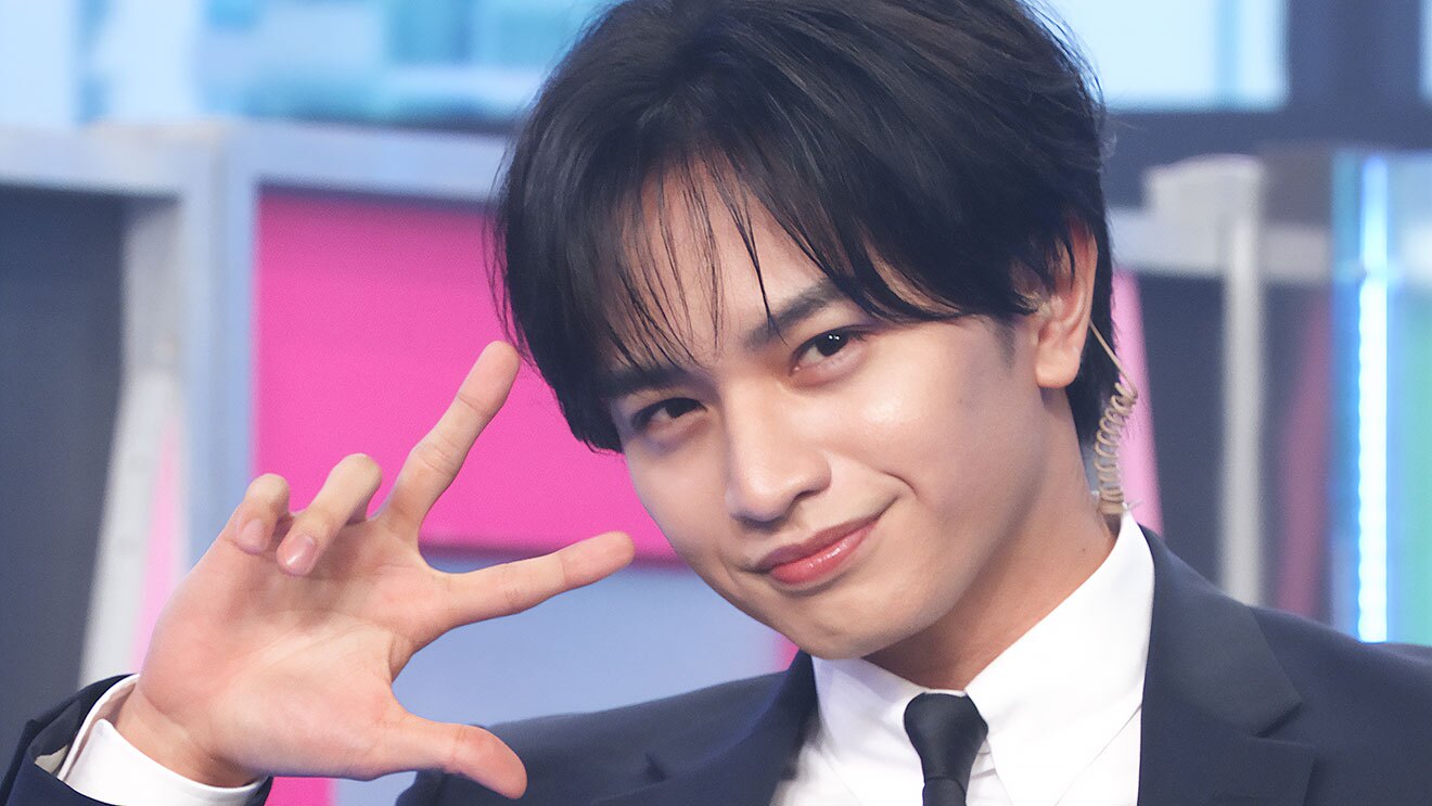 撮影スタジオのセットを背景に、顔の横で右手の指3本を立てる男性のアップ。黒いスーツで黒いネクタイ、耳のうしろにコードが伸びている。