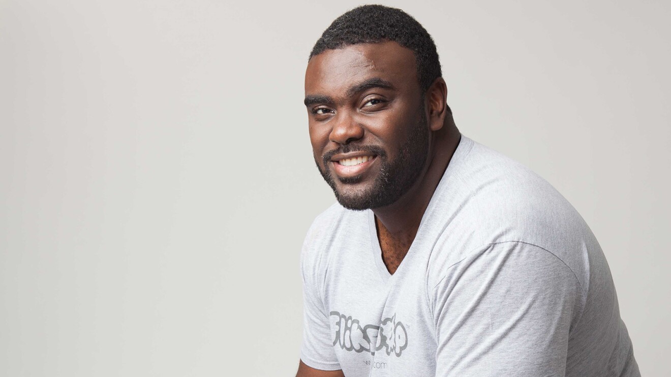 An image of a man smiling for a photo in front of a white background. His shirt says "Flikshop."