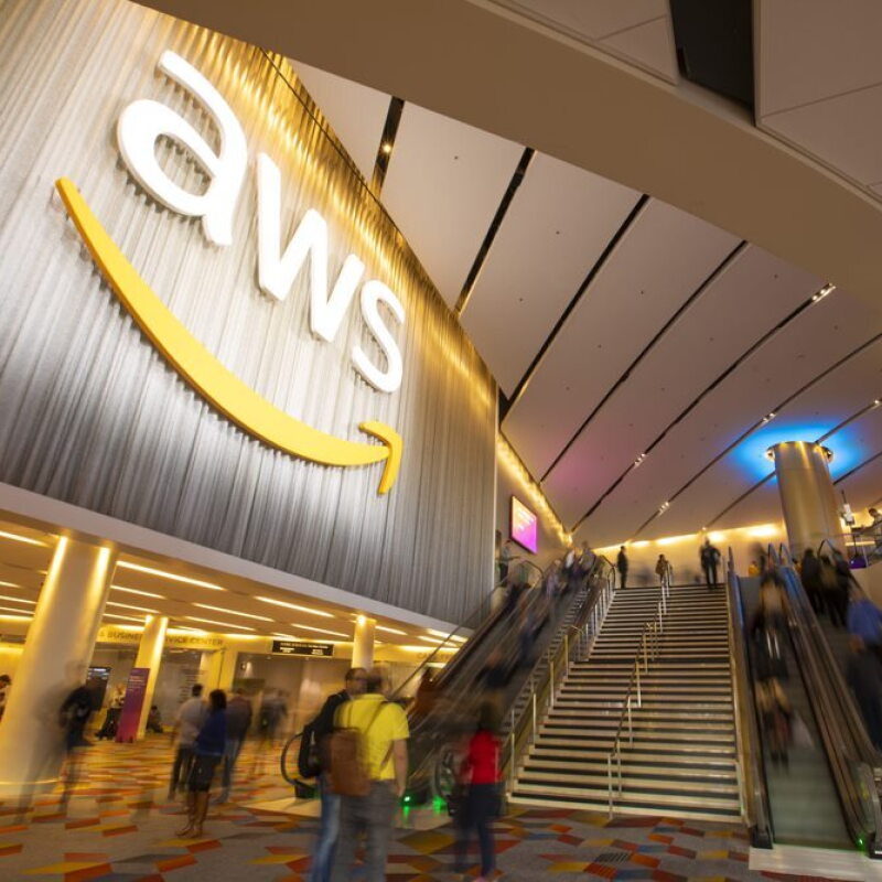 Edificio_AWS. Entrada al edificio de AWS de Amazon. En la entrada con el suelo gris y rombos de color rojo, naranja y amarill. Hay cuatro escaleras automáticas y unas escaleras en medio. El texto es blanco y en una columna del fondo hay luz indirecta azul. Hay gente subiendo y bajando pero difuminada.  