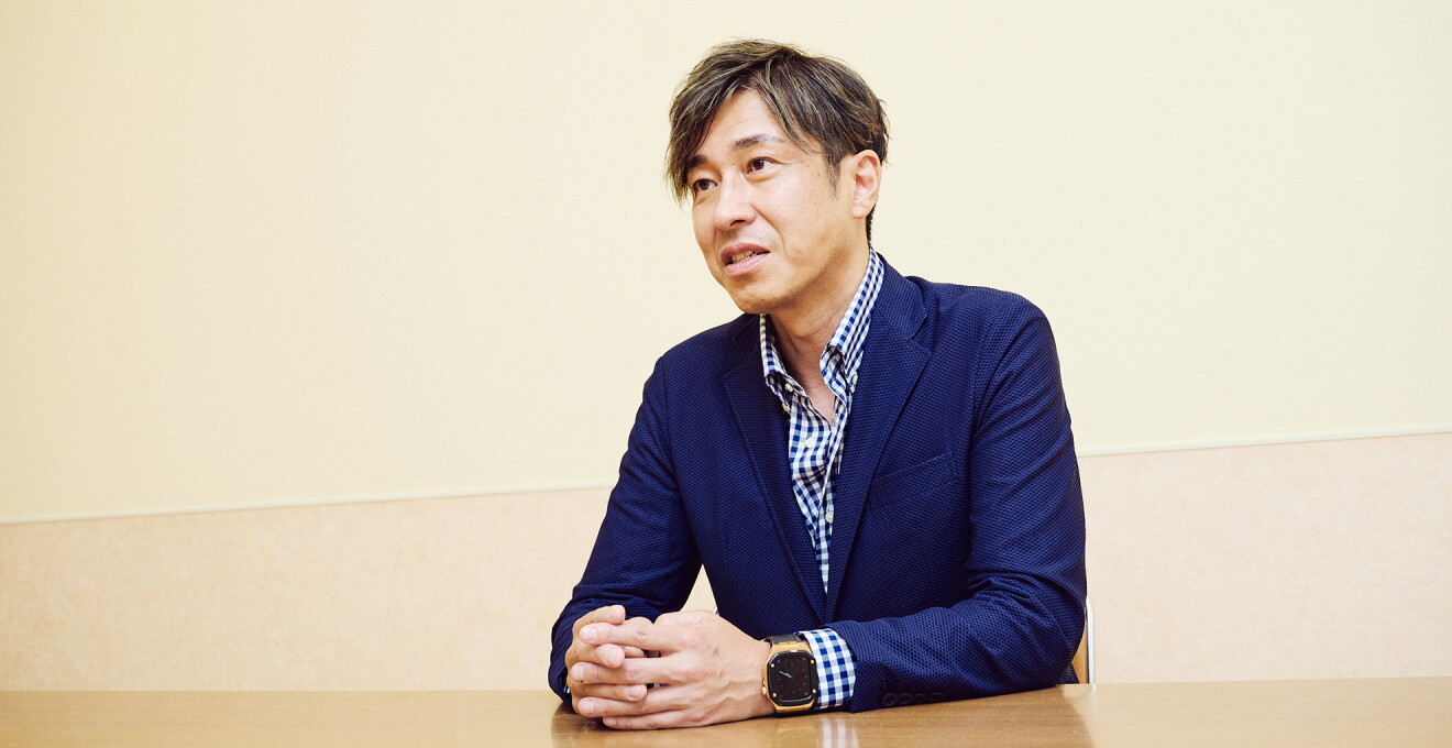 会議室のような場所で椅子に座り話をしている紺色のカジュアルスーツ姿の男性