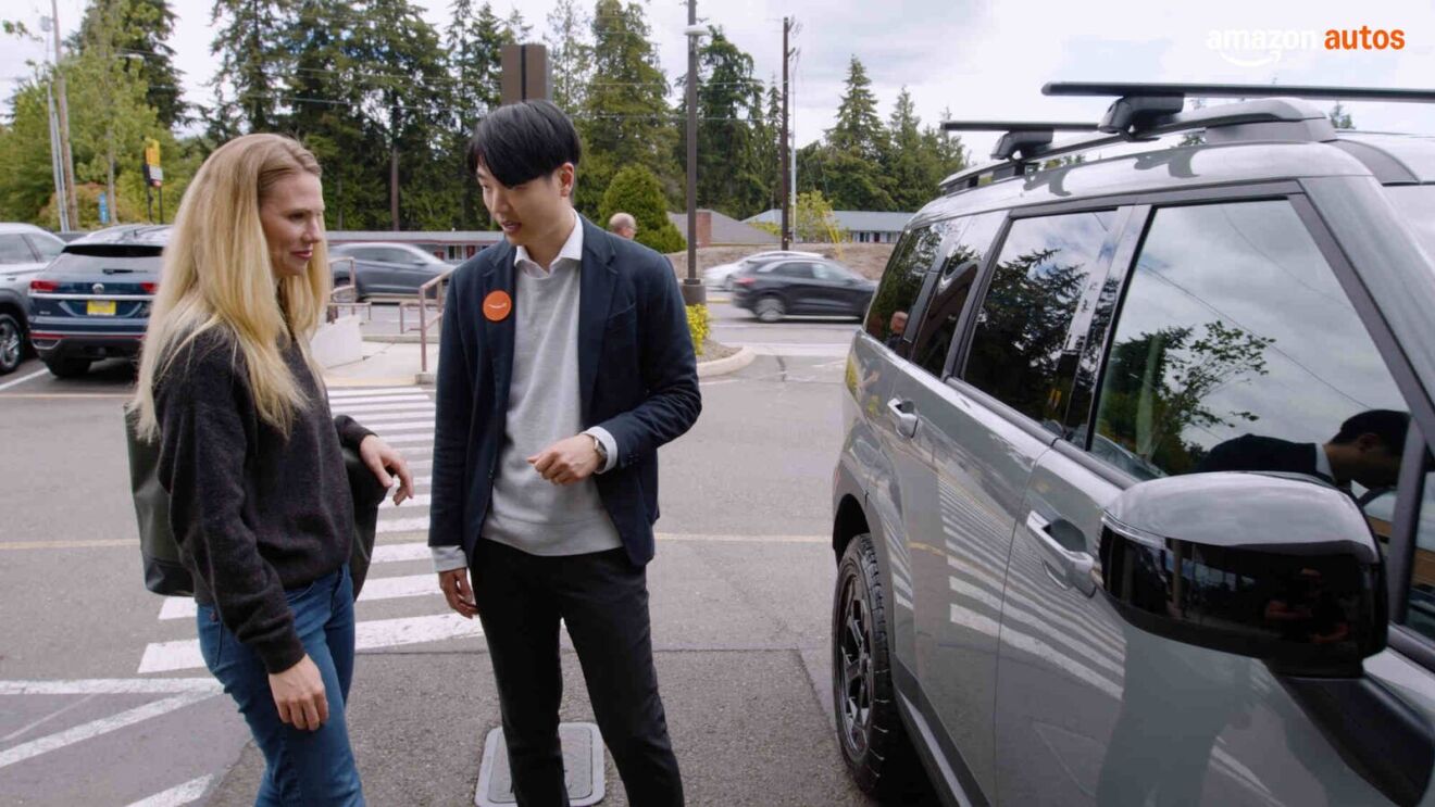 An person wearing an Amazon badge talks to a person looking at a new car in a parking lot.