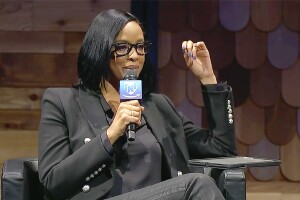 An image of Amina Hussein sitting on a panel discussion and holding a microphone with a "TNF" logo on it.  