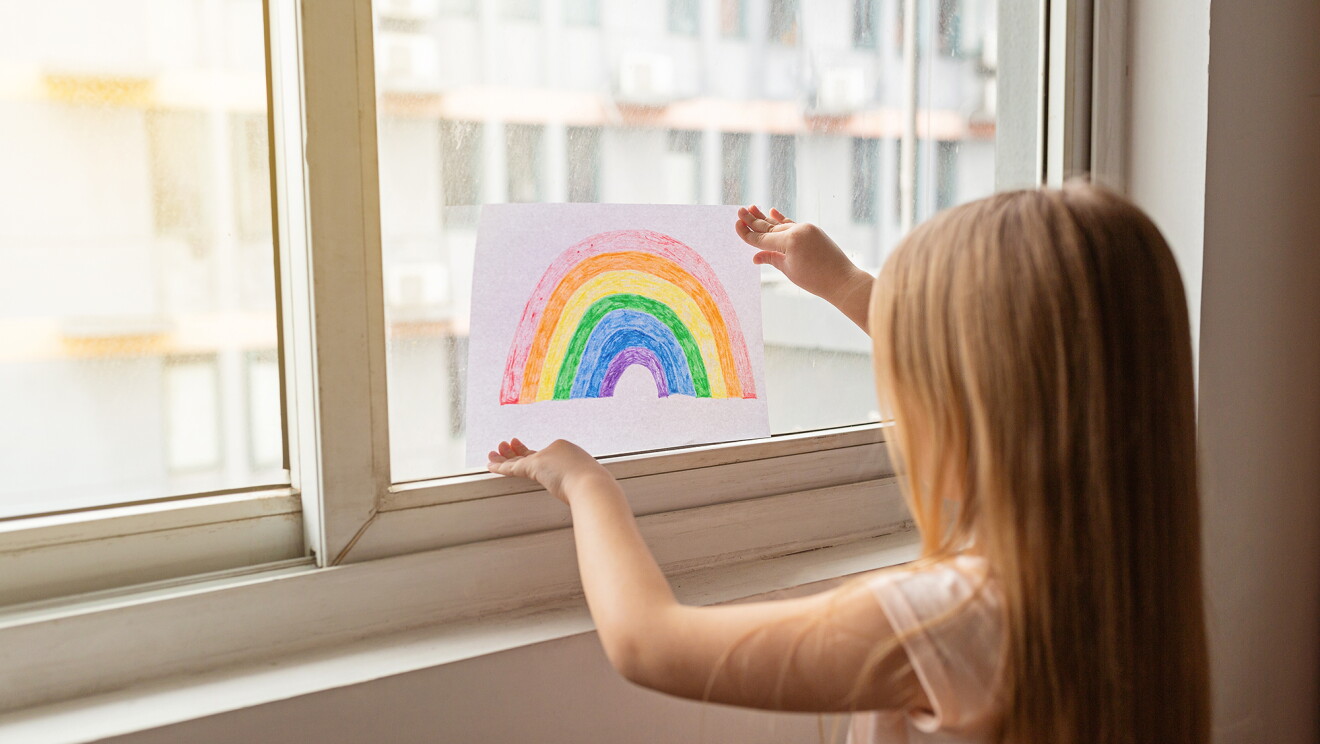 A child placing homemade rainbows in the window of their home, during COVID-19 pandemic.