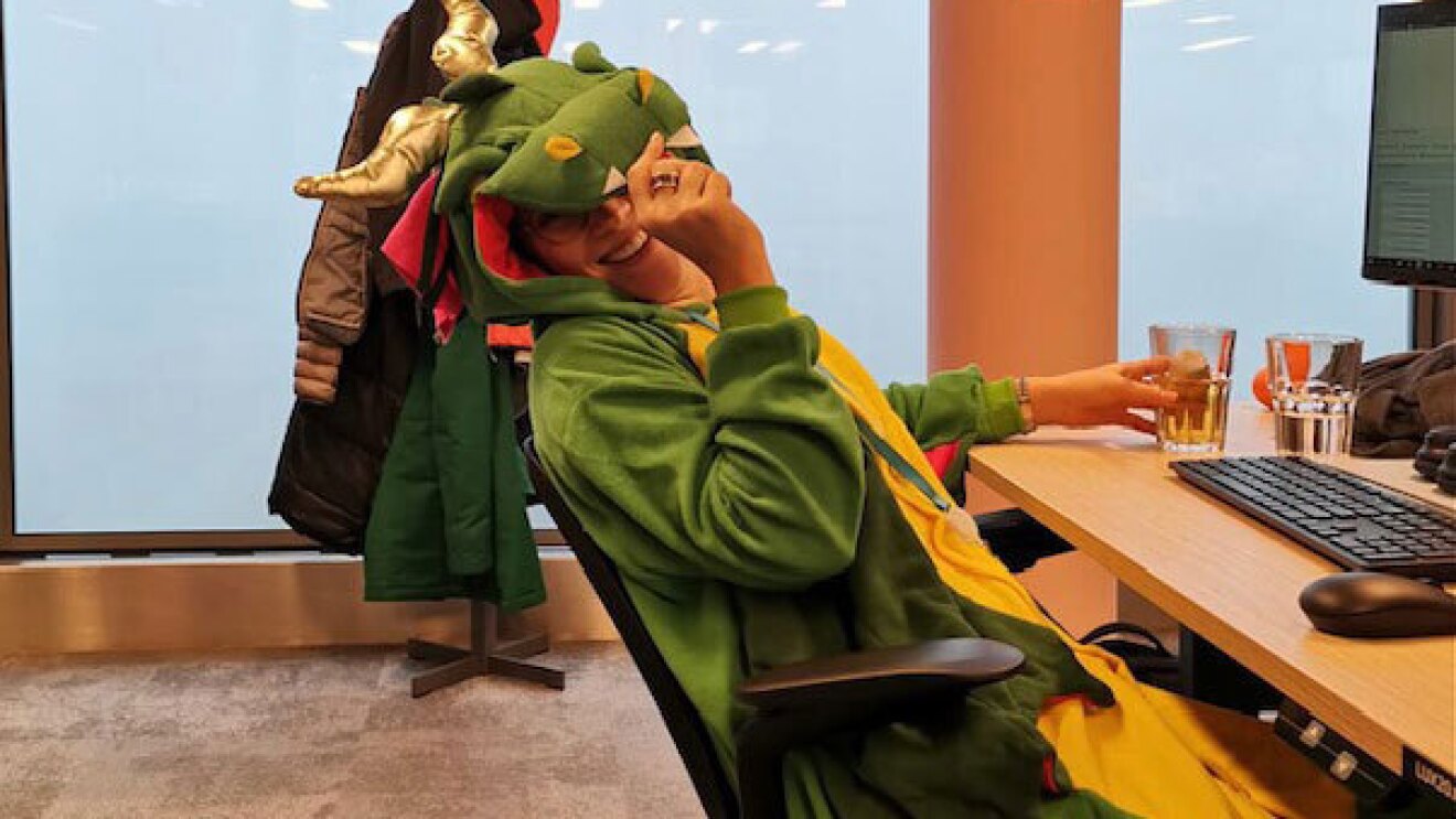 An image of Antonia sitting at her desk at her AWS office smiling while wearing a dinosaur costume.