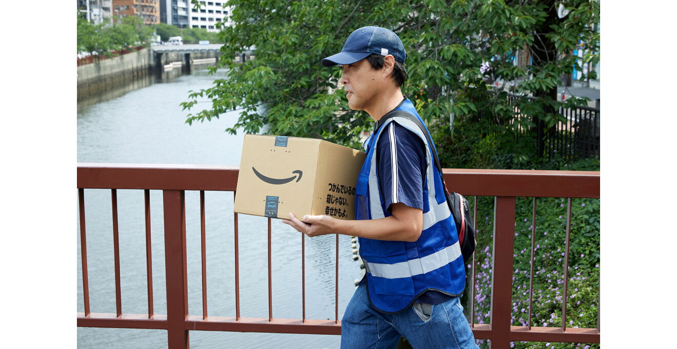 街中の川にかかった橋の上を、荷物を抱えて歩く人物。キャップをかぶり、作業用ベストを着用。箱の表面にはAmazonのロゴがある