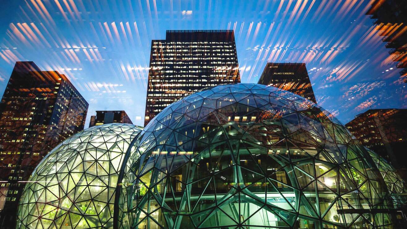 An image of the outside of Amazon's offices at the center of its Seattle headquarters. The camera is looking up at the buildings and the Spheres from the ground. There is a seagull flying in front of the buildings and blue skies in the background.