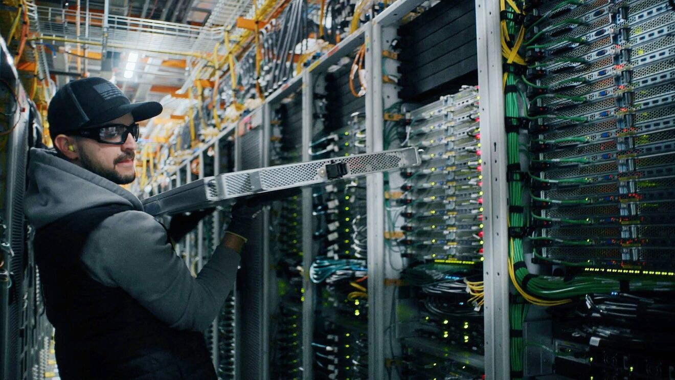 A man wearing a black cap, goggle, and grey jacket is removing a part from the computer setups.