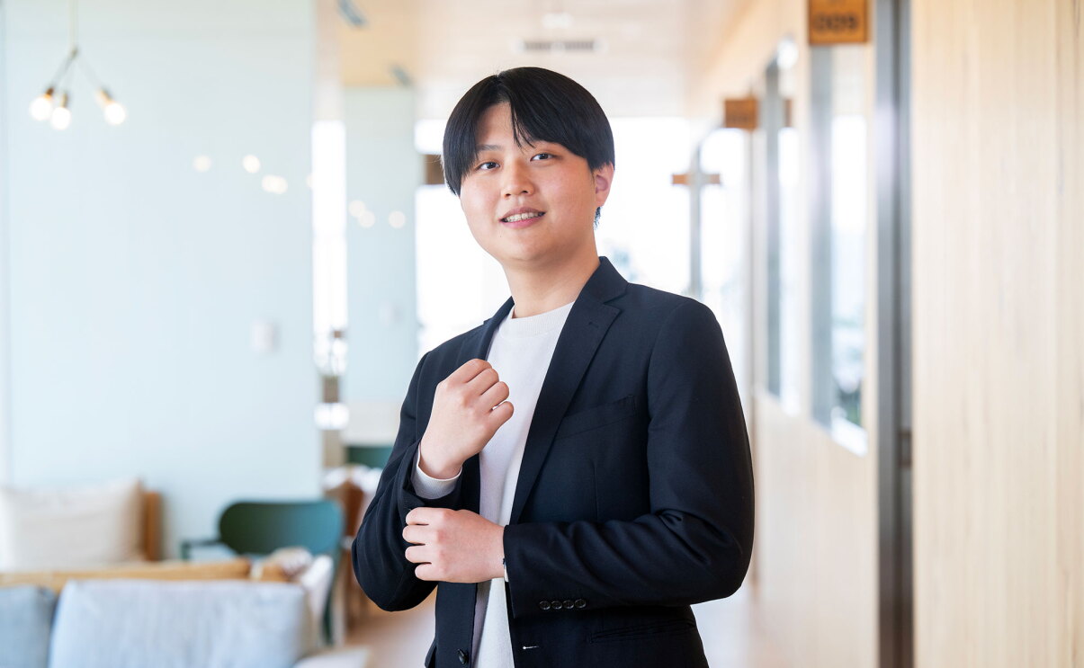 明るいオフィスの廊下で、ネイビージャケットと白のトップスを着用した男性が立っている。廊下の左側にソファが並び、右壁面には複数のドアとプレートが配置されている。