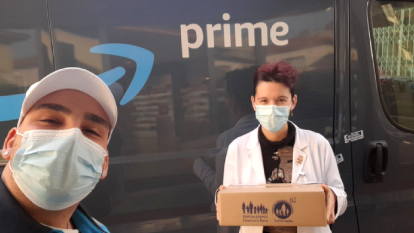 Un autista Amazon consegna un pacco di medicinali ad una farmacista, di fronte ad un furgone con il logo Prime. 