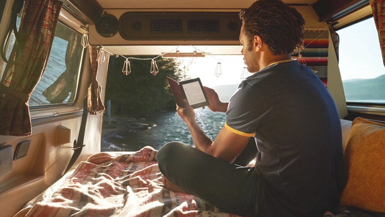 A man reading from a Kindle Paperwhite sits in the back of a camper van, in front of a large body of water.
