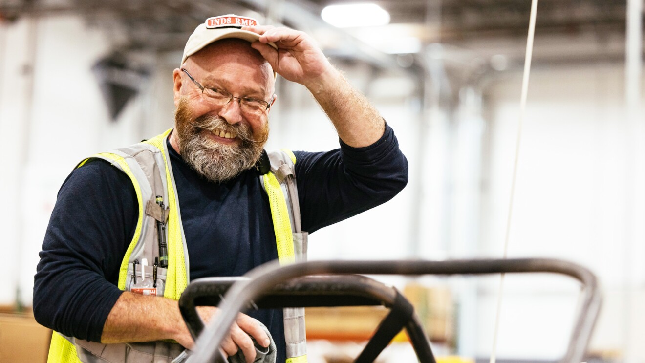 An operations employees wears a yello safety vest as he lifts up his hat and smiles.