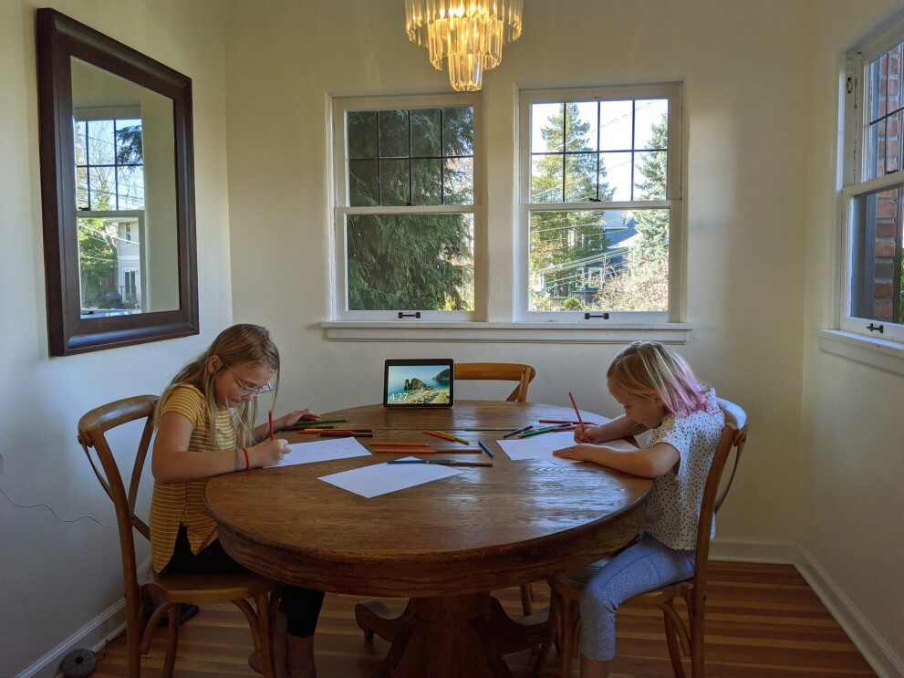 Girls sit at a dining room table and use colored pencils to draw.