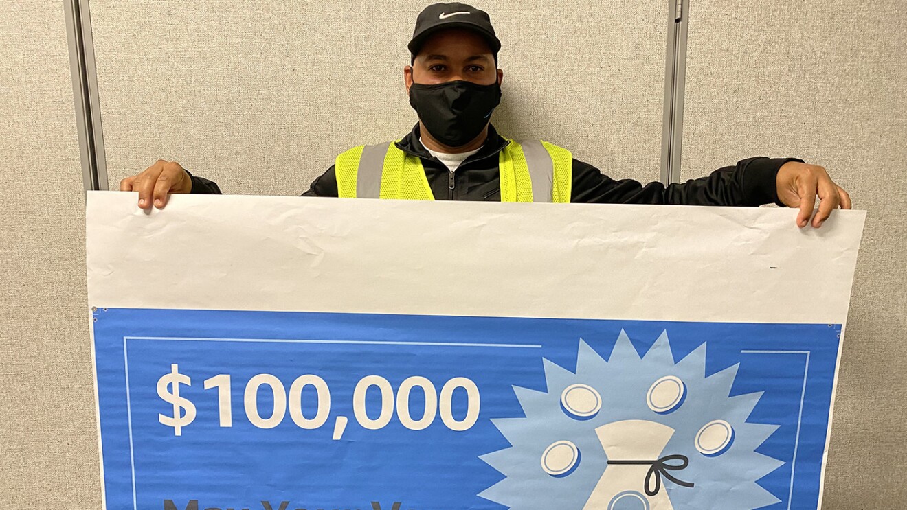 An image of a man holding a large check made out for $100,000 that says "Max Your Vax Winner." He is wearing a mask and a yellow safety vest.