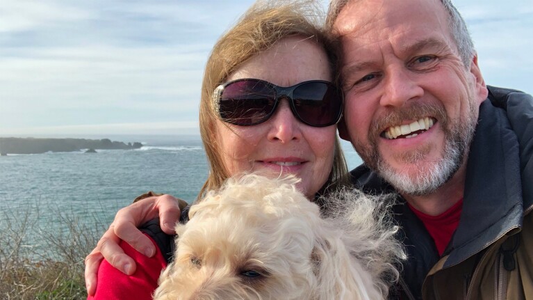 David, Cynthia and their dog smiling at the camera with the ocean in the background.
