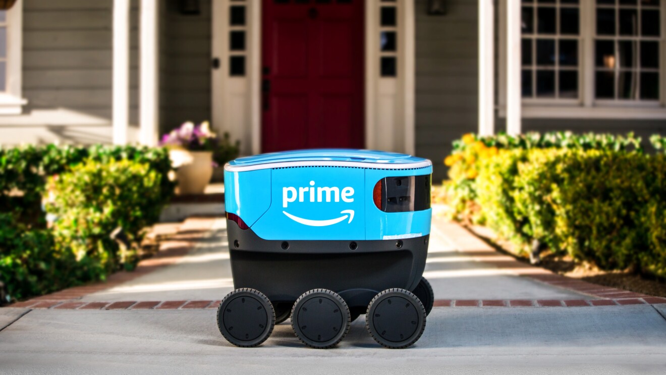 A bright blue, sleek, six-wheeled Amazon Scout delivery robot parked on the concrete walkway of an American home 