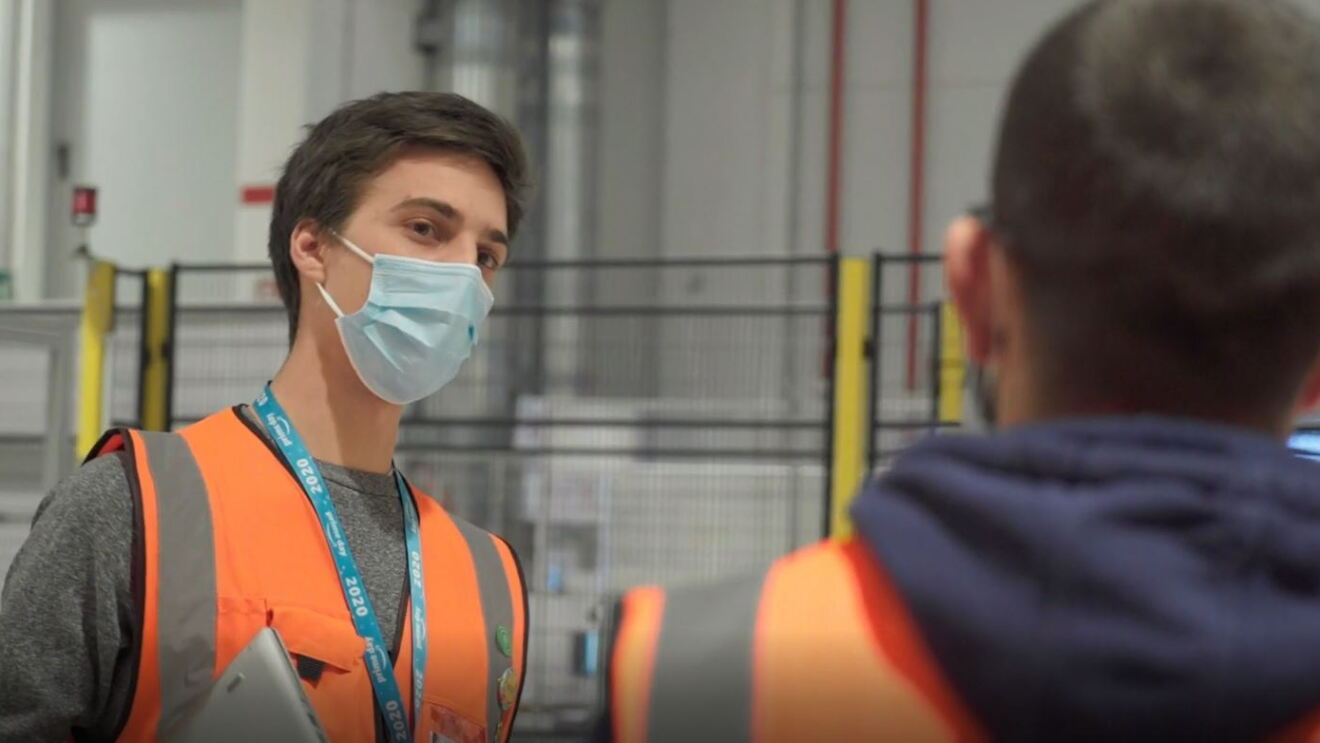 Dipendente Amazon in un magazzino che chiacchiera con un suo collega, girato di spalle. Il dipendente indossa una mascherina chirurgica e un giubbino catarinfrangente, è un uomo con i capelli scuri e corti. 