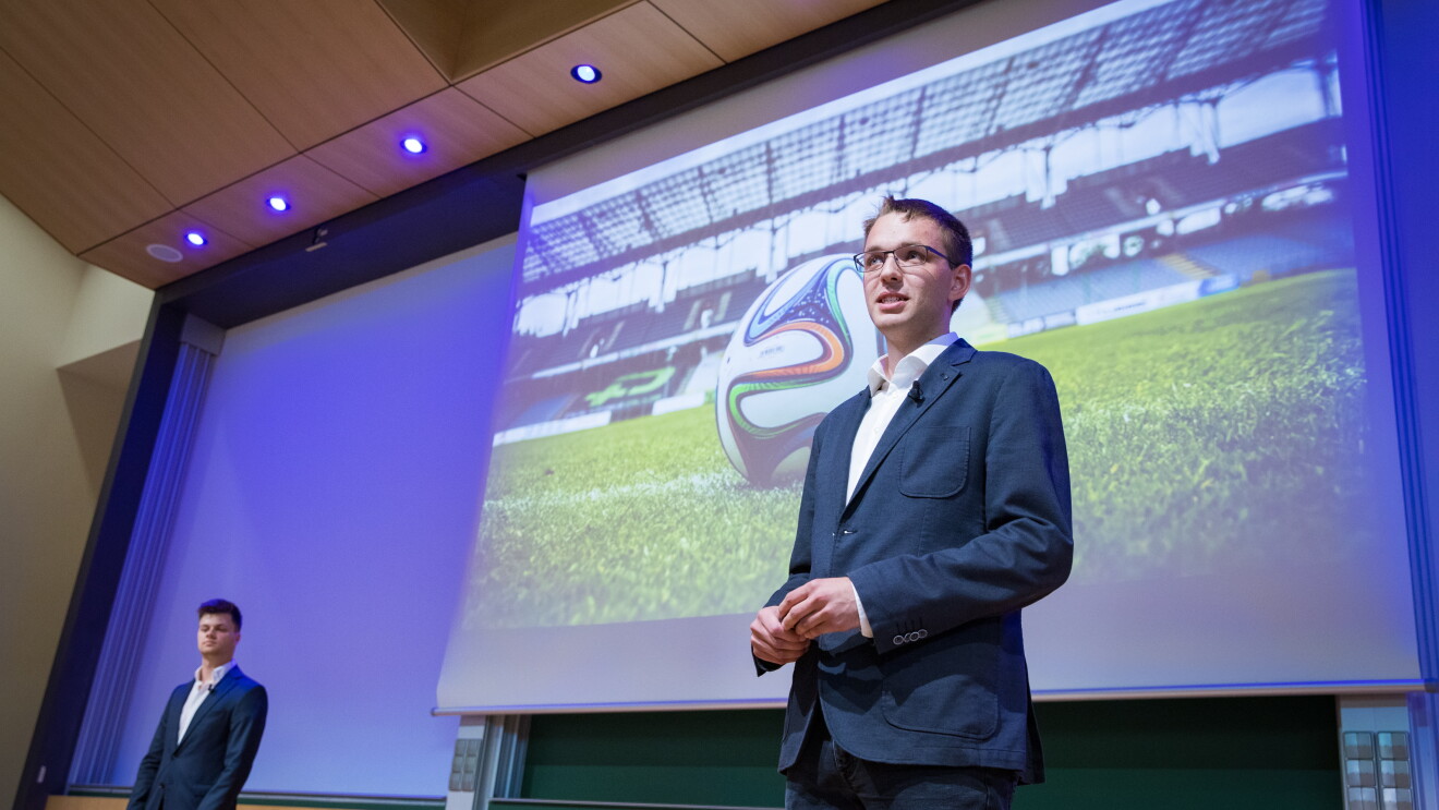 Zwei junge Männer halten einen Vortrag, im Hintergrund ist eine Präsentation zu sehen