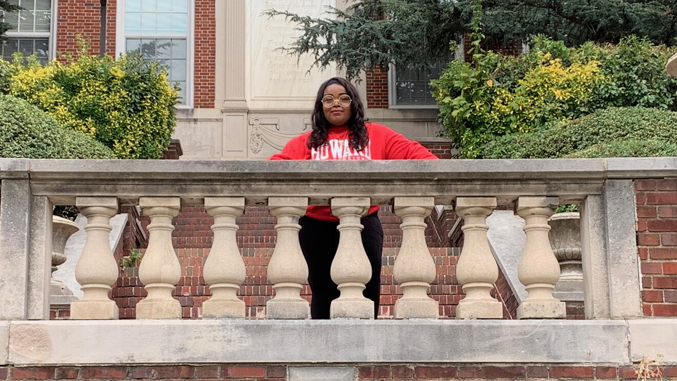 A student photographed on a college campus. She wears a red sweatshirt with the word "Howard" in white letters.