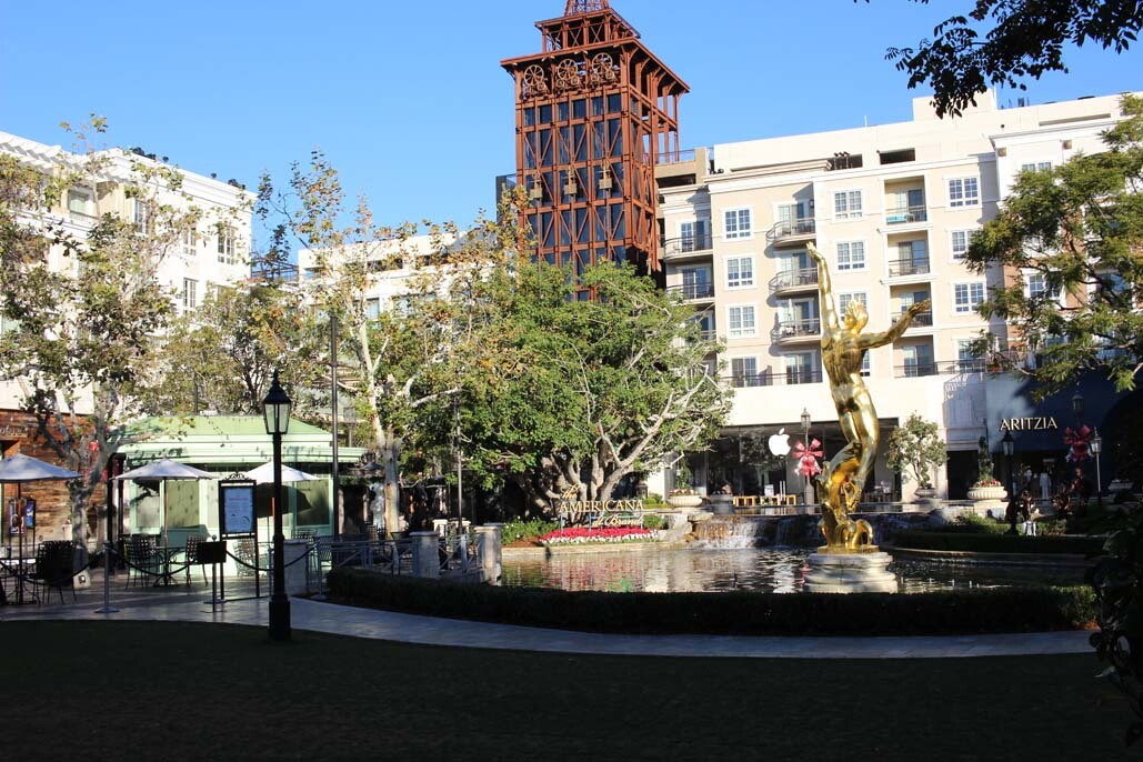 An image of a large fountain with a gold statue of a man in the middle. The fountain is in the middle of a mall with stores surrounding it.