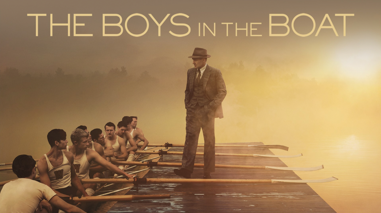 A man in brown suit is standing on a bay staring at boat with rowing team full of young men.