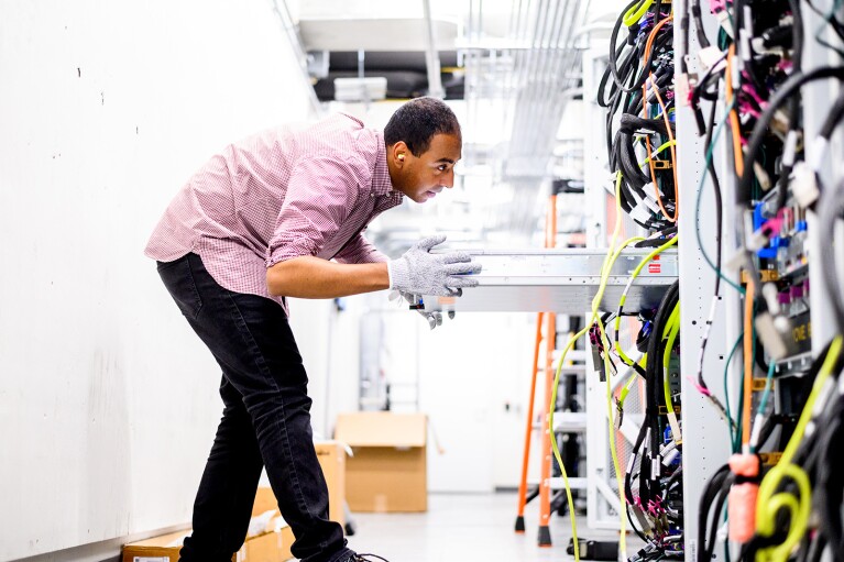 An image of a man pulling a tray out of a wall in the AWS chip lab. The wall has a bunch of wires along it. 