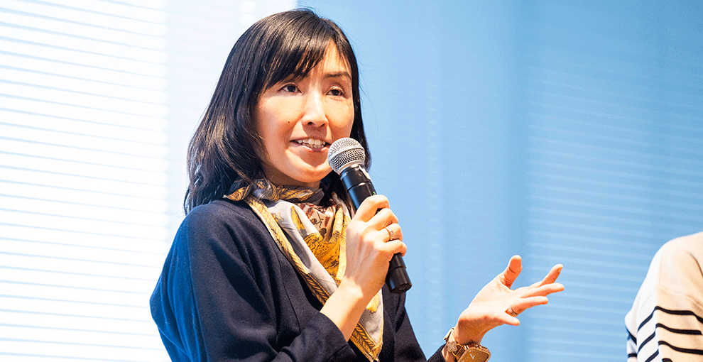 マイクを右手で持った女性。女性の背後にはブラインド。