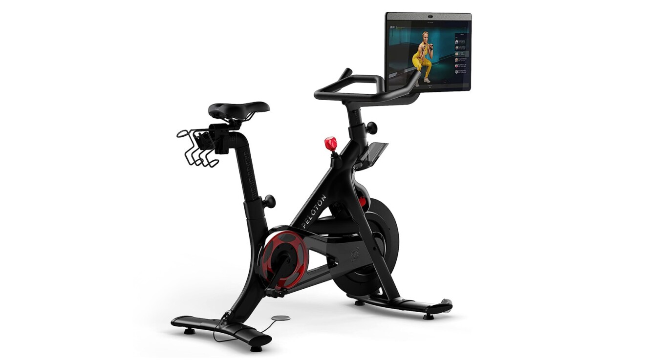 An image of a black Peloton bike with red details. There is a screen on the handles that shows an instructor demonstrating a squat exercise.