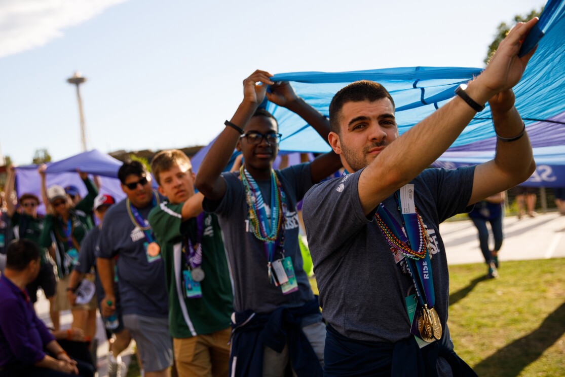 Special Olympics athletes are seen in the foreground and background, as they carry the ceremonial Special Olympics flag during the closing ceremony. Many of the athletes are wearing medals from the games, in the photo.