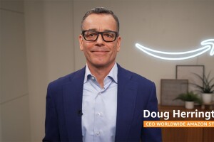 Doug Herrington wears a navy suit and stands in front of a lit up Amazon smile logo hanging on the wall.