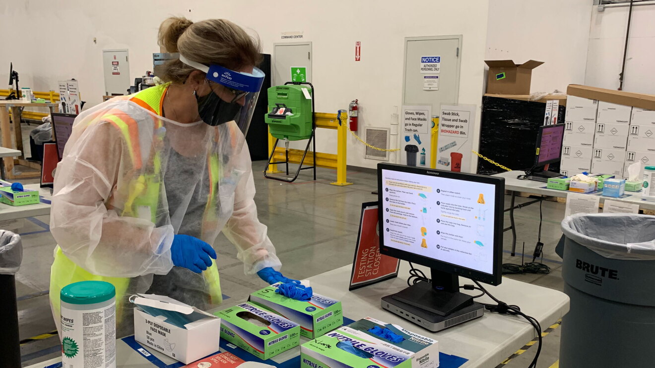 A woman in full COVID-19 personal protective wear and a safety vest stands next to a table that has COVID-19 testing supplies and a computer monitor.