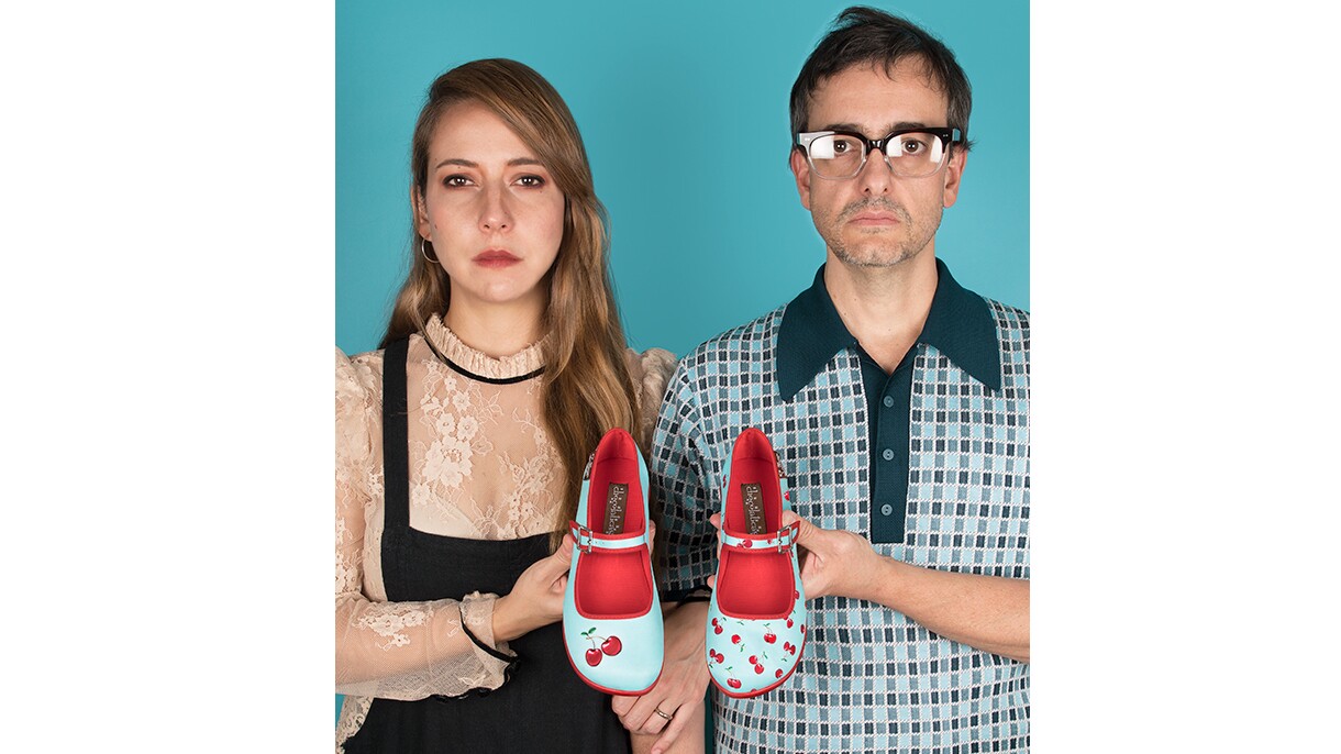 An image of a man and a woman standing in front of a teal background for a photo. They are each holding on shoe in a pair of colorful Mary Jane sandals. The shoes are light blue/teal with red inlining and red cherries on them. Both shoes look slightly different from one another.