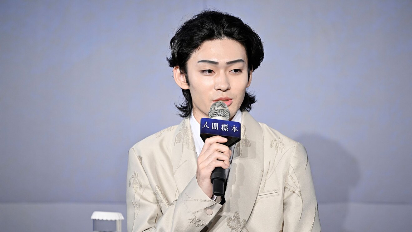 グレーの壁を背景に、マイクを持って話す男性(市川染五郎さん)。ベージュのスーツに白シャツを着用。右手に持ったマイクには「人間標本」と書かれたパーツが付いている。スーツのジャケットは同じ色で植物の刺しゅう入り。