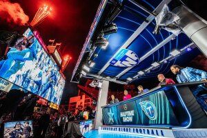 An image of the Thursday Night Football hosts sitting at the announcing table on the field. There is a large, blue screen in front of them and red fireworks taking flight in the air above.