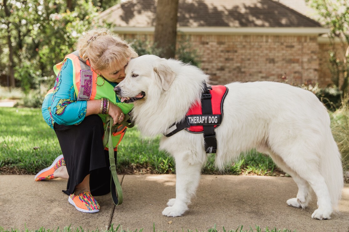 A woman in a bedazzled Amazon Flex vest hugs a white dog, wearing a therapy dog service animal vest.
