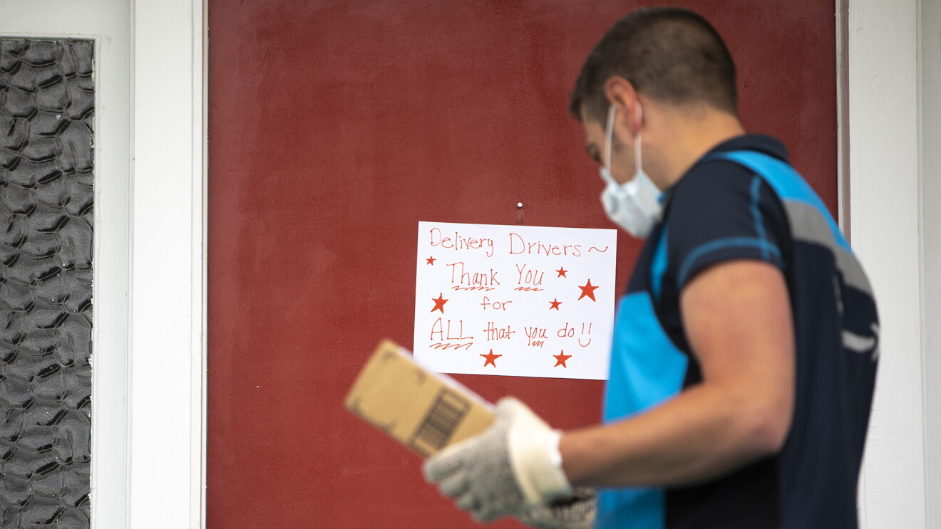 Amazon delivery driver stands in front of a door, to deliver a package. On the door is a sign that says "delivery drivers, thank you for ALL that you do."