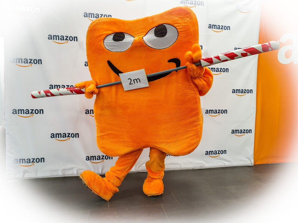 Eine Person im Kostüm des Amazon Maskottchens Pecci Pecci steht auf dem Logistik-Außengelände. Neben ihm hängt an einem Zaun ein Plakat mit dem Hinweis, 2 Meter Abstand zu halten.
